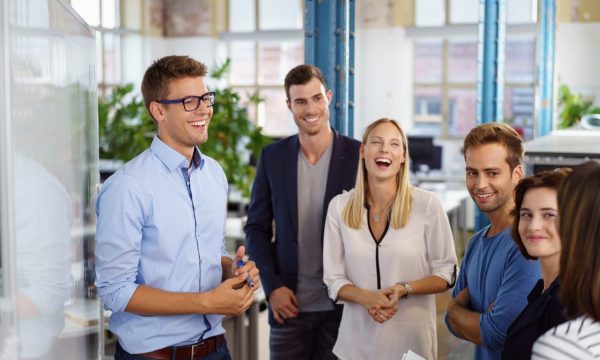 Laughing white collar workers standing around white board during meeting in small office